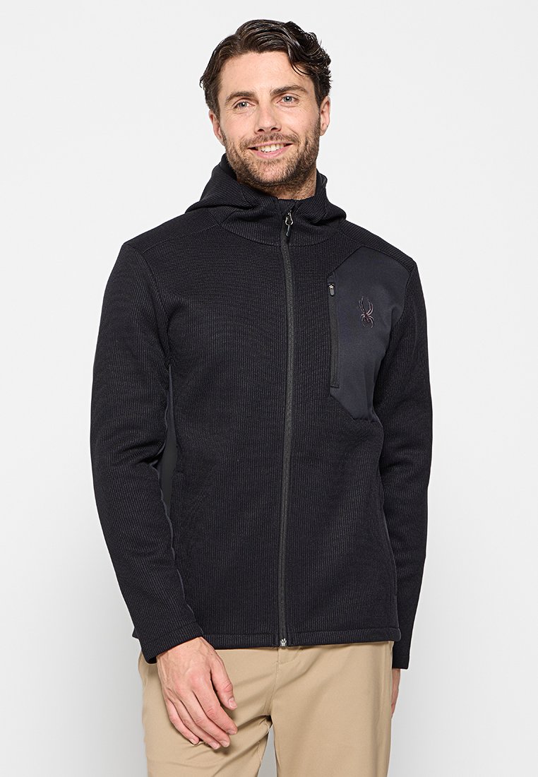 Schwarze Fleecejacke mit gerippter Textur, voller Reißverschluss, verstellbare Kapuze und einer mit Reißverschluss versehenen Brusttasche, die auf der linken Seite ein Logo trägt.