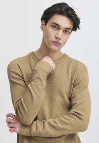Beige strikkegenser med rund hals, ribbestrikkede mansjetter og en myk tekstur. Modellen står med armene krysset foran en nøytral bakgrunn.