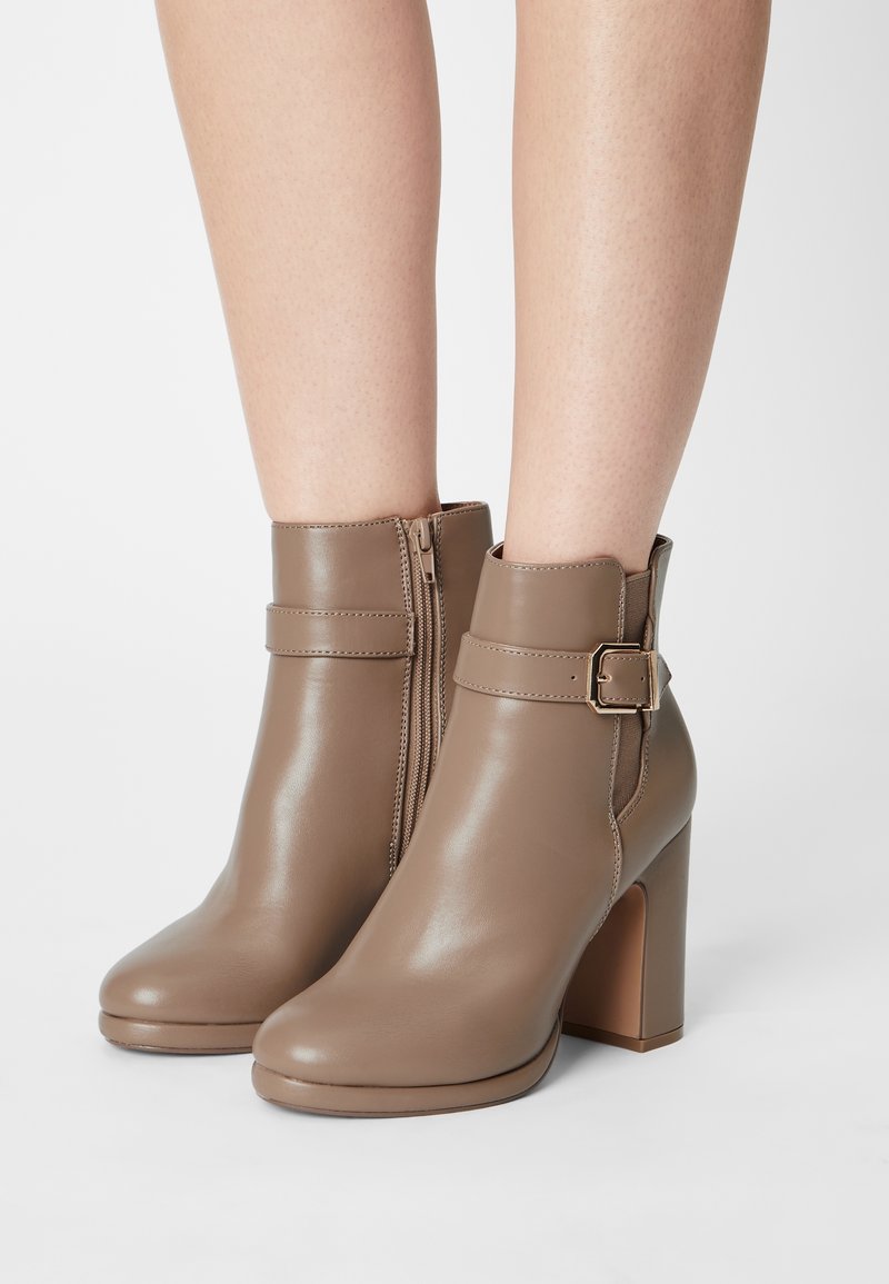 Botas de tobillo de color topo lisas con tacones cuadrados, cremalleras laterales y correas decorativas con hebillas, llevadas sobre piernas al desnudo.