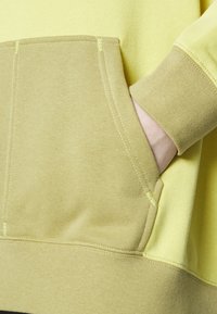 Felpa gialla con tasca e polsini color beige, realizzata in un tessuto morbido con cuciture visibili e una texture liscia.