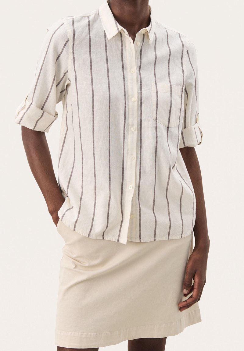 Personne portant une chemise blanche à rayures verticales marron avec manches retroussées, associée à une jupe beige longueur genou.