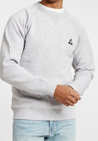 Uomo che indossa un maglione grigio chiaro a collo rotondo con un piccolo logo triangolare nero sul petto e jeans azzurri chiari, mentre sistema la manica.