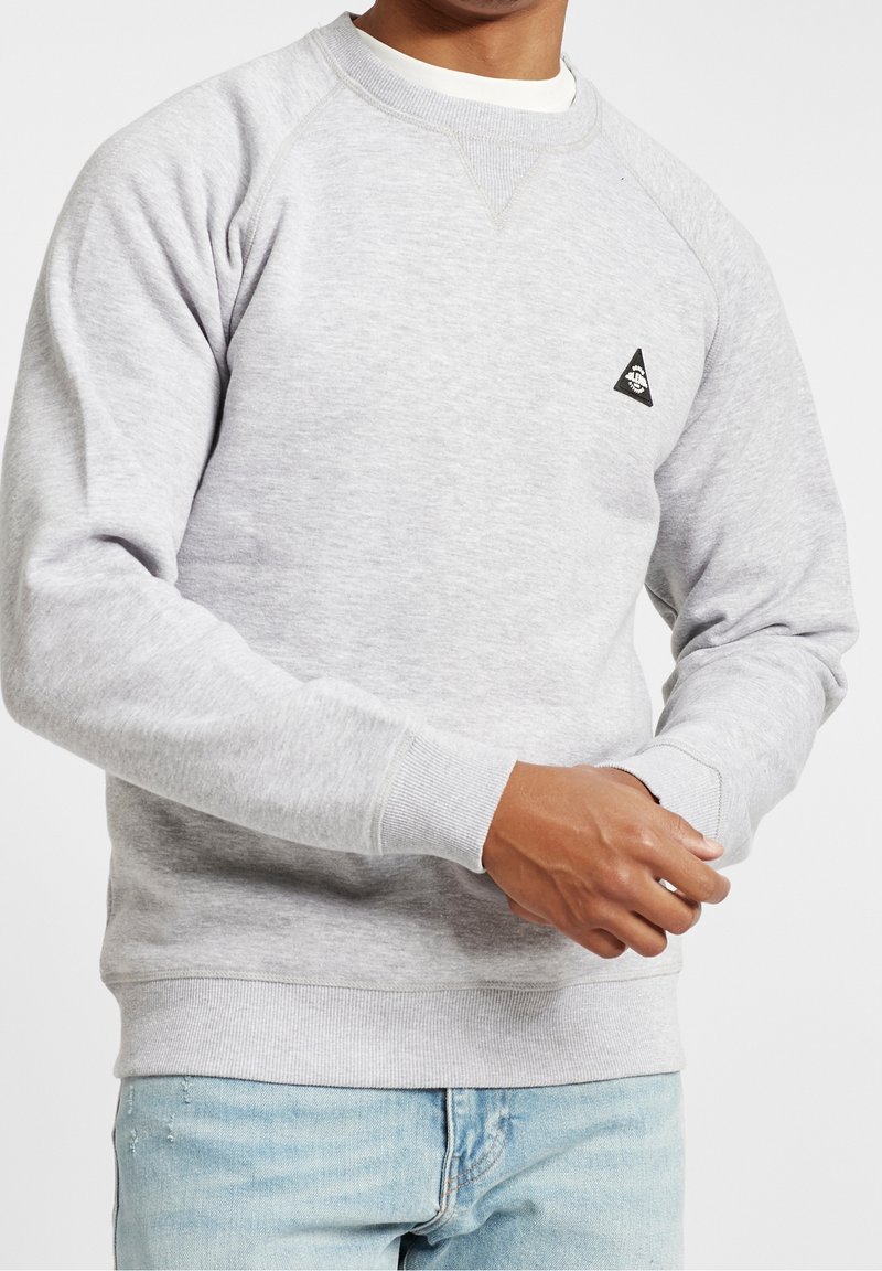 Uomo che indossa un maglione grigio chiaro a collo rotondo con un piccolo logo triangolare nero sul petto e jeans azzurri chiari, mentre sistema la manica.