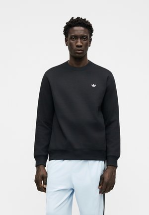 Muž nosící černou mikinu Adidas s bílým logem trefoil na hrudi a světle modré kalhoty s černými pruhy, stojící proti jednoduchému pozadí.