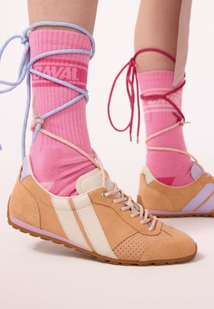Pieds portant des baskets beige et blanches avec des chaussettes roses, chacun enveloppé et attaché avec des lacets de différentes couleurs autour des chevilles.