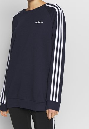 Marineblauwe Adidas sweater met witte drie-strepen details op de mouwen en klein logo op de borst, gedragen met zwarte broek met witte zijstrepen.