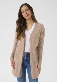 Cardigan beige en maille douce, ouvert sur le devant avec manches longues, associé à un t-shirt blanc et un jean bleu. Design simple et coupe décontractée.