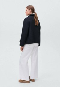 Femme avec des cheveux longs attachés en arrière, portant une veste noire, un pantalon large blanc et des chaussures brunes, se tenant devant un fond uni.