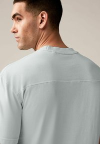 Tricou din bumbac gri deschis, cu guler rotund, mâneci scurte și o detaliere a cusăturii pe spate. Textură moale și netedă.