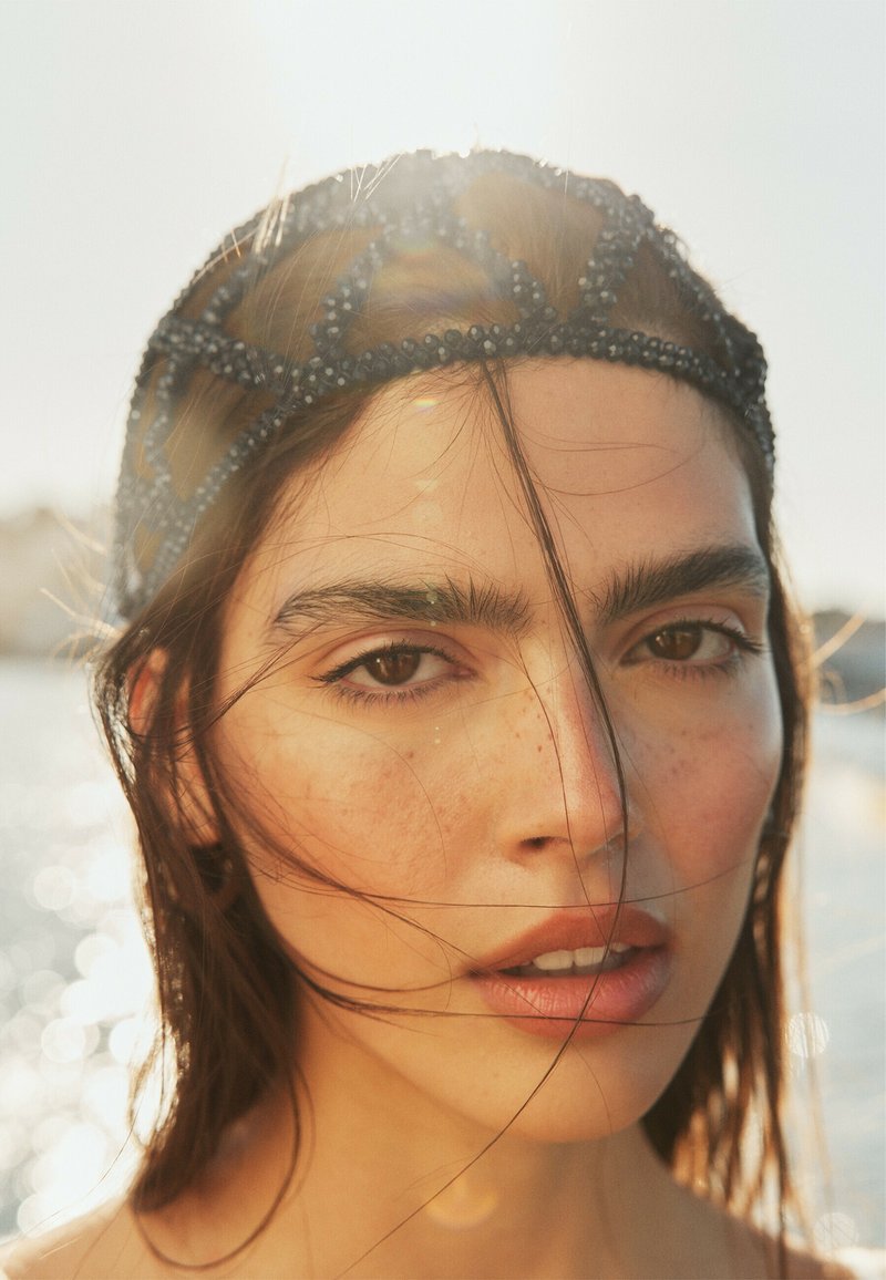 Gros plan d'une femme à la peau éclairée par le soleil, aux cheveux foncés détachés, et portant un bandeau noir orné de perles, en extérieur près de l'eau, à la lumière naturelle douce.