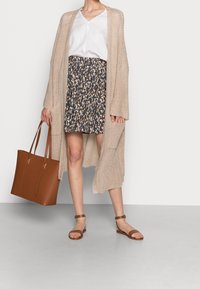 Cardigan lavorato a maglia beige indossato sopra una blusa bianca e una gonna scura a fantasia. Una borsa a spalla marrone e sandali completano l'outfit. Design semplice e informale.