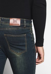 Vaqueros de mezclilla oscura con un acabado desgastado, que cuentan con bolsillos traseros y una etiqueta de cuero marrón grabada con una mariposa y la frase "GOODFORNOTHING."