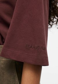 Bordeaux kleurige gemarmerde top met korte mouwen en geborduurd 'ROUGE EDIT' detail op de manchetten. De stof lijkt zacht en iets gestructureerd.