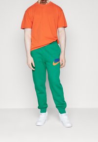 Oranžové bavlněné tričko s kulatým výstřihem, doplněné zelenými tepláky z fleecu s modro-oranžským logem Nike. Bílé tenisky.