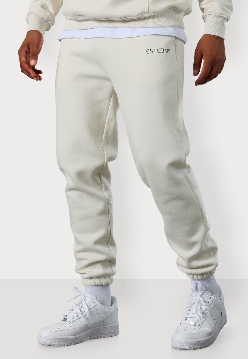 Weiße Jogginghosen aus Baumwolle mit elastischen Bündchen, ausgestattet mit einer Vordertasche und dem gestickten "ESTEEM"-Logo, kombiniert mit weißen Turnschuhen.