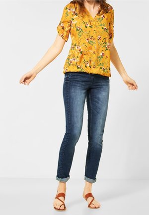 Femme portant un chemisier jaune fleuri, un jean bleu foncé retroussé et des sandales marron à lanières, debout devant un fond clair uni.