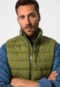 Grön puffig väst med hög krage, dragkedja fram och sömdetaljer. Bärs över en blå denimskjorta med långa ärmar och knapplåtar.