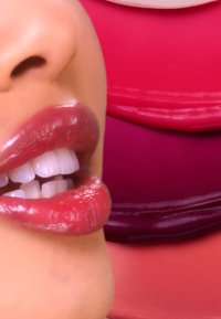 Close-up van glanzende, licht getinte lippen met een gladde, glanzende textuur, tegen een vervaagde achtergrond van gelaagde roze lippenstiftkleuren.