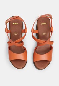 Narančaste kožne sandale s debelim potplatima, s križnim trakama i kopčom oko gležnja. Unutrašnjost prikazuje oznaku, "Bata Made in Italy."