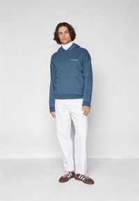 Sudadera con capucha azul y bolsillo frontal, llevada sobre un cuello de tortuga blanco, combinada con pantalones blancos y zapatos marrones. Conjunto casual.