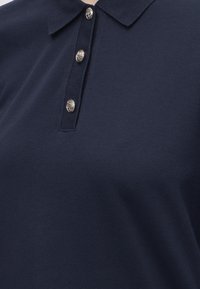 Polo bleu foncé en tissu texturé avec un col, présentant une rangée verticale de trois boutons décoratifs en argent.