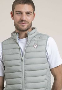 Gilet rembourré gris clair avec col haut, zip sur le devant et patch logo. Le tissu semble lisse avec une texture douce. Porté par-dessus un t-shirt blanc.