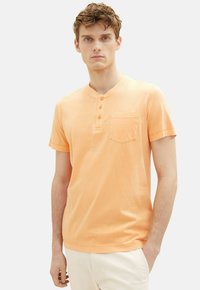 Ferskenfarvet kortærmet henley skjorte med tre-knaps placket, brystlomme og blød stoftekstur. Model står foran en neutral baggrund.