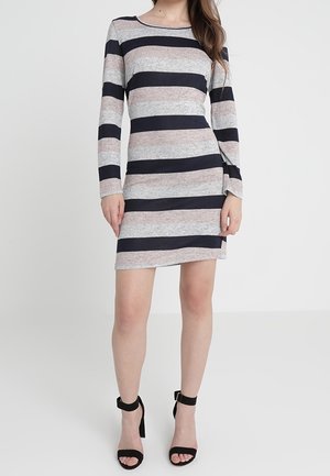Kvinde iført en langærmet kjole, der går til knæene, med brede vandrette striber i sort, grå og beige kombineret med sorte højhælede sandaler.