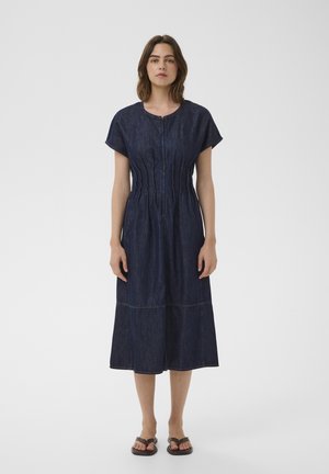 Kobieta stoi przodem, ubrana w ciemnoniebieską, krótkorękawową sukienkę z denimu o długości do połowy łydki z suwakiem z przodu oraz czarne sandały.