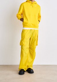 Geel hoodie met koord, en bijpassende gele cargobroek. Getextureerde stof, hoge fit, twee zijzakken, zwarte instappers.