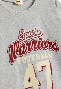 Grijze katoenen sweatshirt met rode en crèmekleurige opdruk: "Sports Warriors Football" en het nummer "47." Ribbelboord bij de hals.