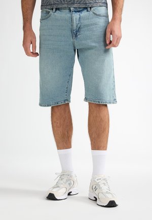 Lichtblauwe denimshorts met een rechte snit, rafelige zoom, vijf zakken en een verweerde afwerking. Draagt witte sokken en sportschoenen.