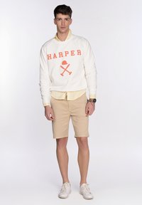 Sudadera blanca con "HARPER" en naranja, superpuesta a una camisa de collar amarilla clara. Unos pantalones cortos beige y unas zapatillas blancas completan el conjunto.