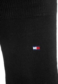 Schwarze Socke mit geripptem Muster und einem kleinen rot-weißen-blauen Logo nahe dem Bündchen. Hergestellt aus einem glatten, dehnbaren Material.