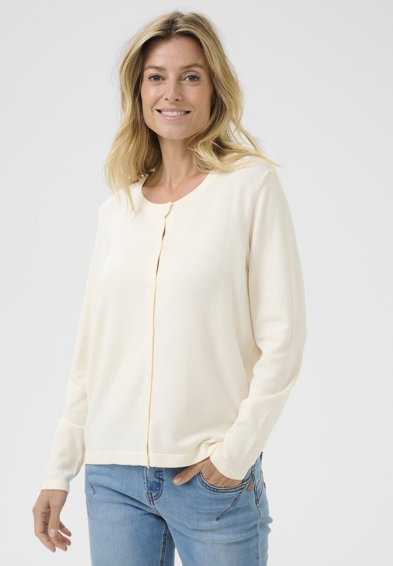 Cardigan crema realizzato in morbido tessuto, con colletto rotondo, chiusura con bottoni e maniche lunghe; abbinato a jeans azzurri chiari.