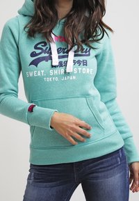 Felpa turchese con lacci bianchi e testo stampato. Presenta una tasca a marsupio frontale e polsini a costine, indossata con jeans blu.