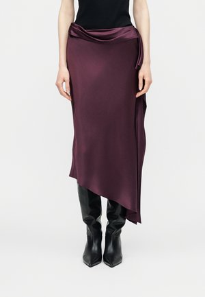 Femeie purtând o fustă midi asimetrică drapată, de culoare violet închis, cu detaliu legat, bluză neagră și cizme negre din piele până la genunchi.