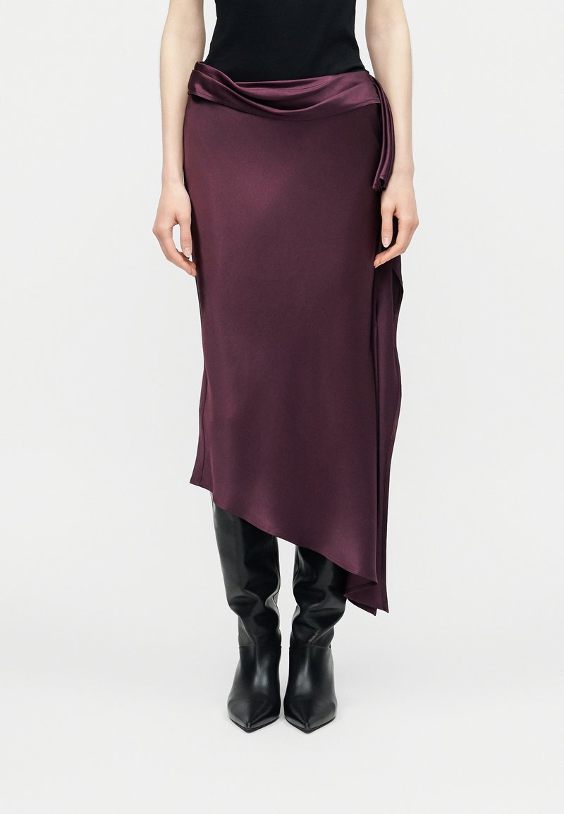 Femeie purtând o fustă midi asimetrică drapată, de culoare violet închis, cu detaliu legat, bluză neagră și cizme negre din piele până la genunchi.