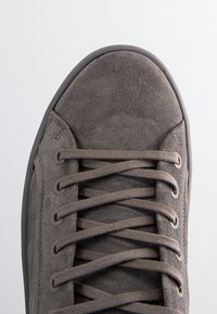 Zapatilla de ante gris con punta redondeada, que presenta cordones planos, ojales de metal y parte superior texturizada. Suela de goma con acabado suave.