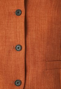 Καφέ ύφασμα σε σχέδιο καρέ με σακάκι που έχει τέσσερις μαύρους κουμπιά. Διαθέτει ίσια γραμμή και λεπτομέρεια με πλαϊκή τσέπη, με λεία υφή.