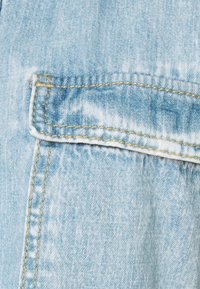 Ljust blå denimtyg med en horisontell ficklock, detaljerad med gul sömmar längs sömmarna.