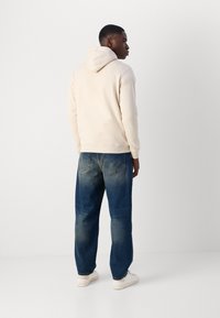 Felpa beige con cappuccio posteriore, abbinata a jeans blu medio a vita larga. Sneakers bianche completano l'outfit. Sfondo semplice.