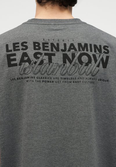 Persoană purtând o cămașă gri cu text negru pe spate, pe care scrie "LES BENJAMINS EAST NOW Istanbul" și un text mai mic dedesubt.
