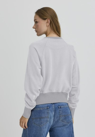 Light gray cropped sweatshirt with ribbed hem and cuffs, featuring a round neckline and subtle branding on the sleeve. Paired with denim jeans.