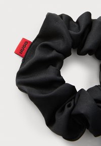 Mustast satäänist valmistatud scrunchie, millel on sile tekstuur ja kokku volditud disain, millel on väike punane silt, millel on kirjas "HUGO". Üldine ringikujuline kuju.