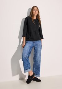 Schwarzes Oberteil mit dreiviertel Ärmeln, lockere blaue Jeans mit hochgekrempelten Säumen und schwarze Slipper. Einfaches Design, bequeme Passform.