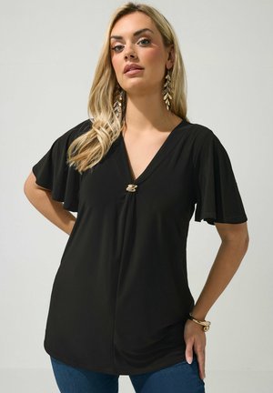 Blonde vrouw met een zwarte top met korte mouwen, V-hals en gouden gesp, gecombineerd met gouden blad oorbellen en een gouden armband.