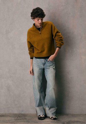 Pullover lavorato a maglia giallo senape con collo a mezza zip, abbinato a jeans blu sbiaditi a gamba larga e scarpe con motivo. Vestibilità rilassata e texture.