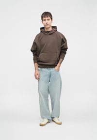 Felpa con cappuccio marrone e tasca frontale, abbinata a jeans larghi blu chiaro e sneakers color crema. Design semplice, vestibilità rilassata.