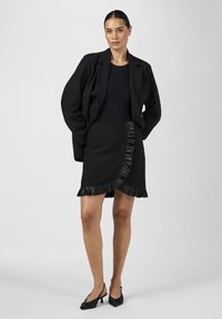 Čierny blazer s prehnanými rukávmi, spárovaný s čiernou mini sukňou s textúrovaným volánkovým lemom a akcentom. Lesklé čierne topánky na podpätku.